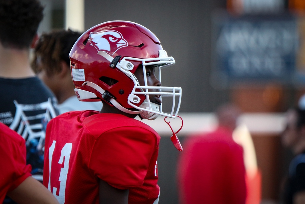 A football player in a red uniform and helmet stands with others, looking to the side.