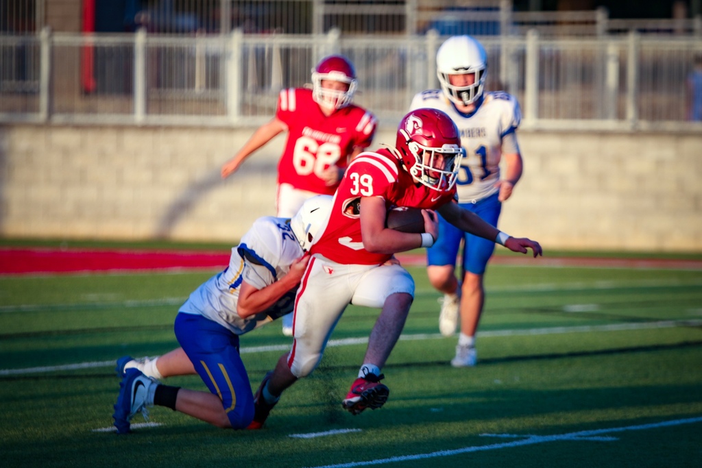 Players in red and blue uniforms compete on a football field; one player in red is tackled by a player in blue.