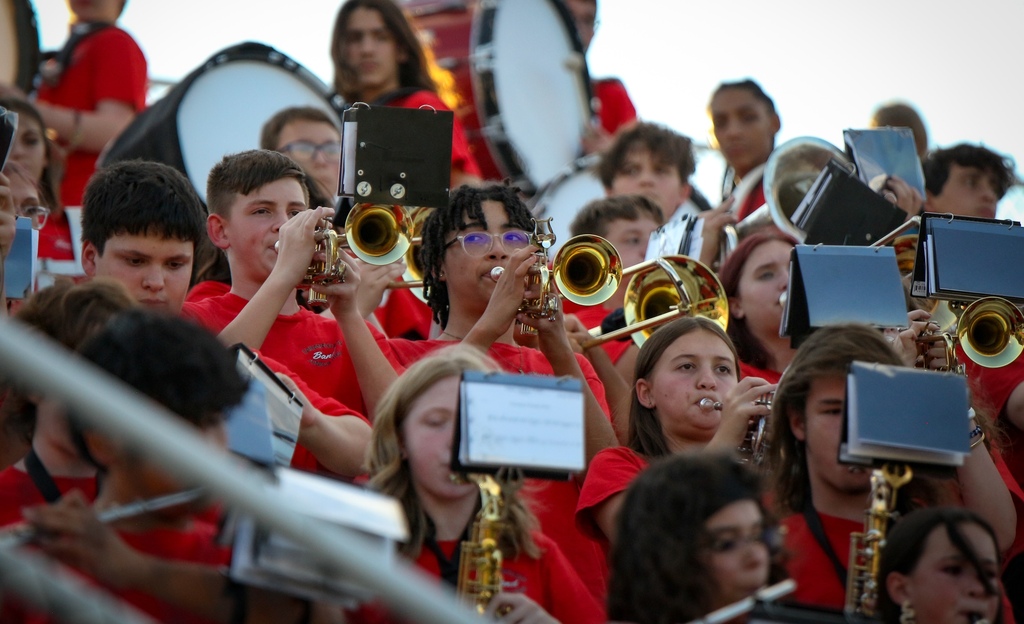A group of people in red shirts play instruments in a stadium. Some play trumpets and saxophones.
