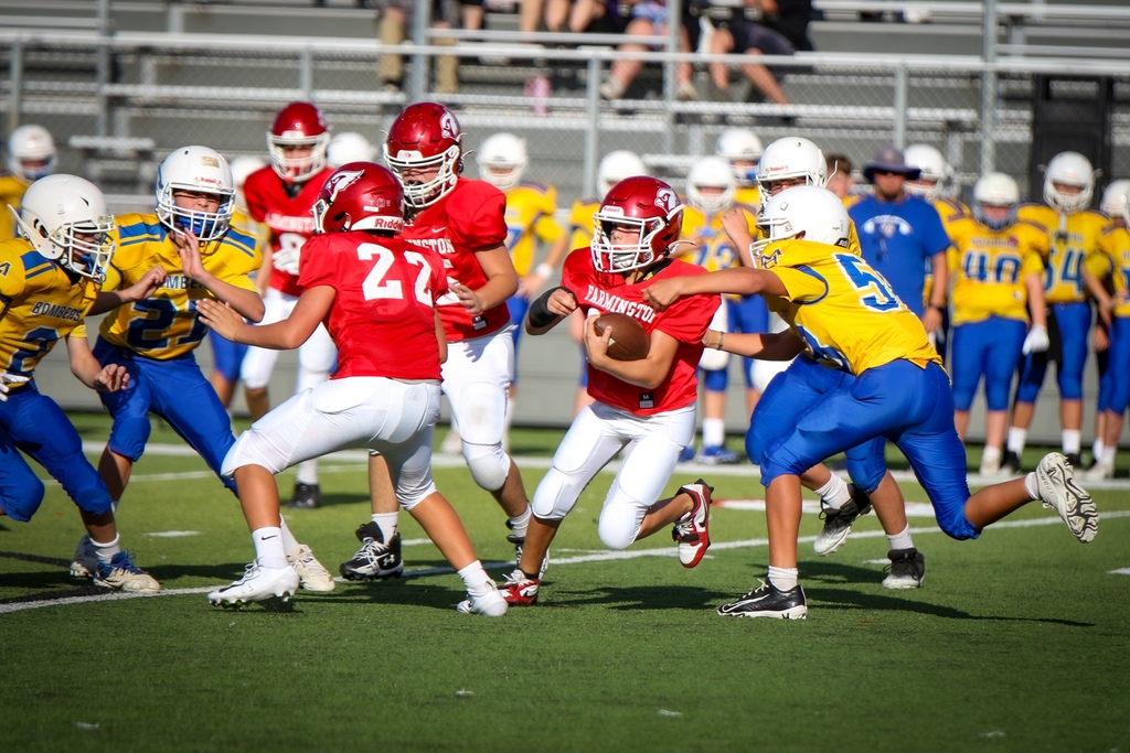 Youth football game on a field. Players in red and blue uniforms compete. A player holds the ball while others tackle.