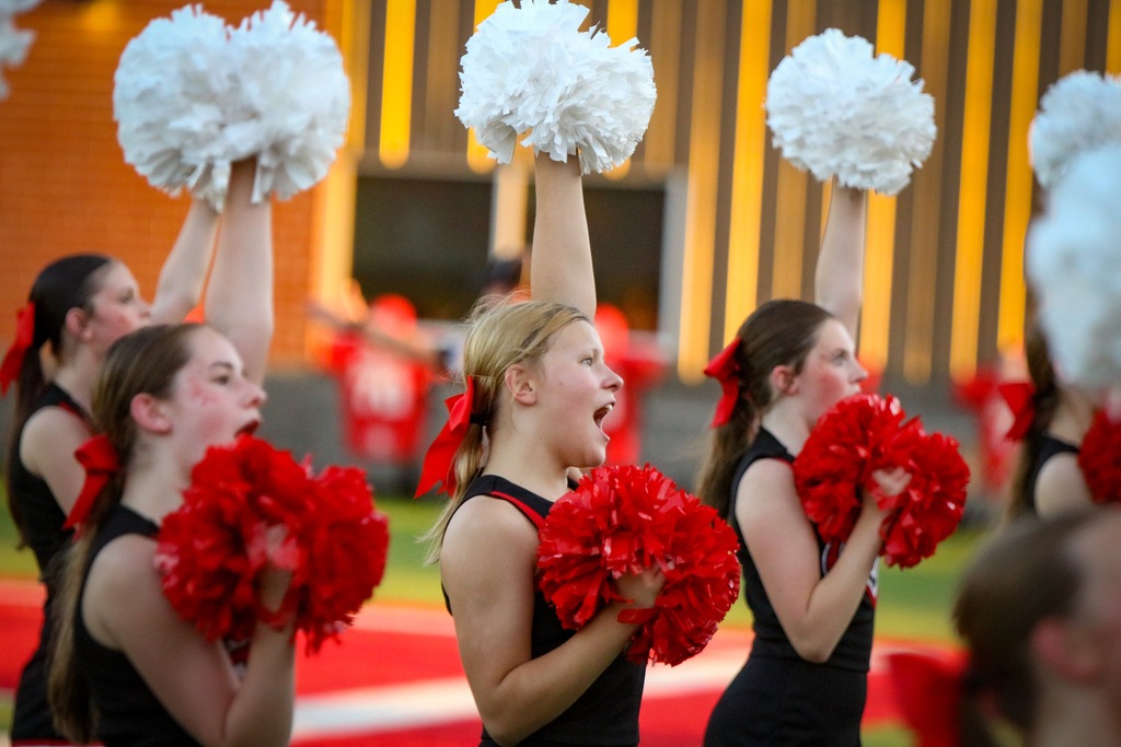 A group of cheerleaders holding red pom poms, cheering in an outdoor sports arena.