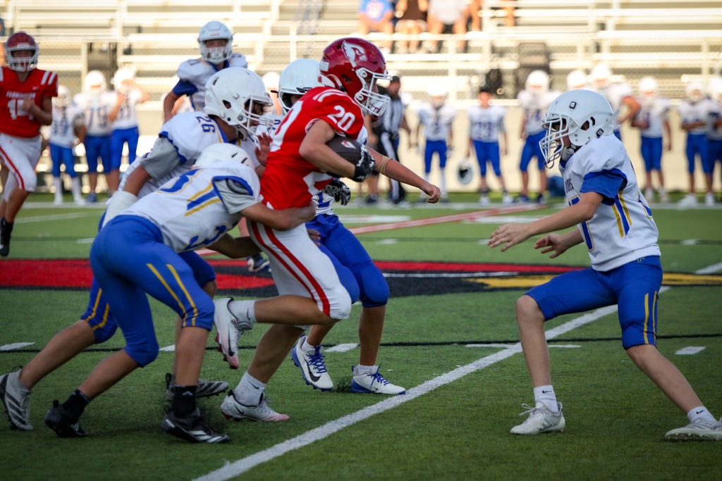 Players in red jerseys clash with those in blue on a football field, with other players and spectators in the background.