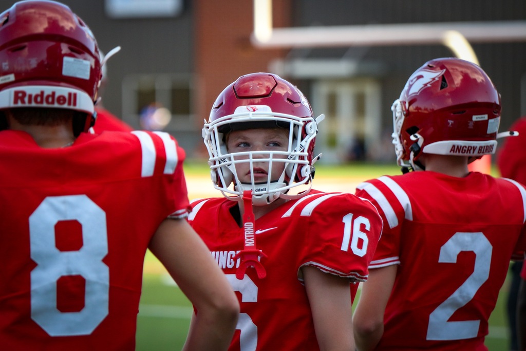 Three players in red jerseys and helmets stand together on a football field. Numbers 8, 16, and 2 are on their jerseys.