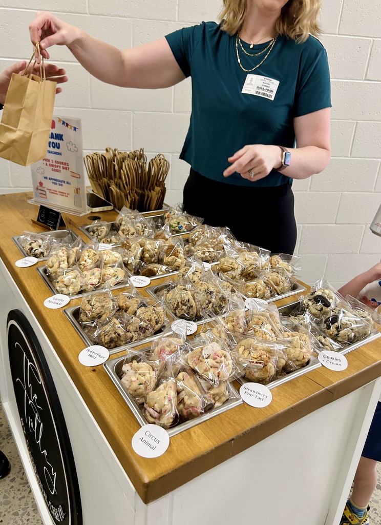 A woman stands at a table with cookies in plastic bags on display. She holds a brown paper bag.