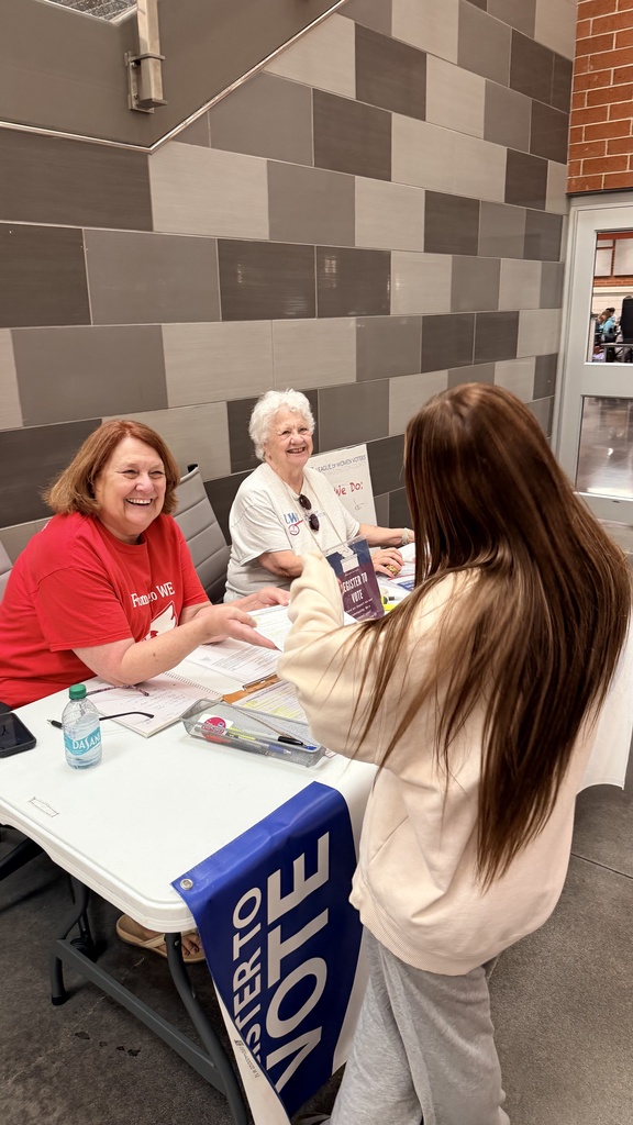 Two women in red shirts seated at a table, one smiling, as another woman in white stands in front of them.
