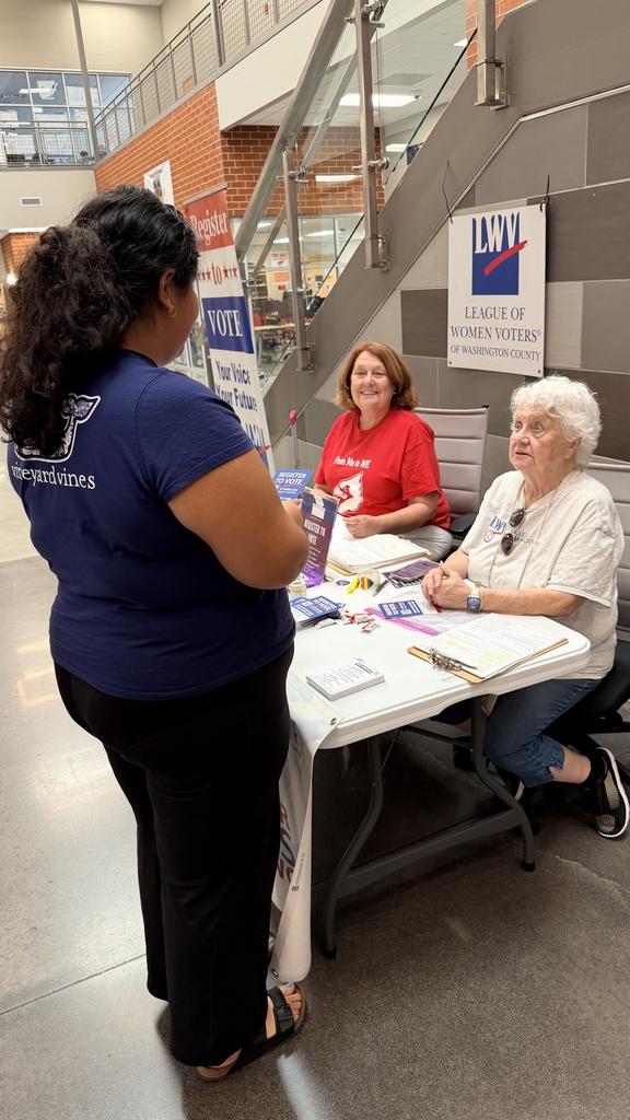 Two women seated at a table with a sign behind them that reads "League of Women Voters." One woman stands facing them.