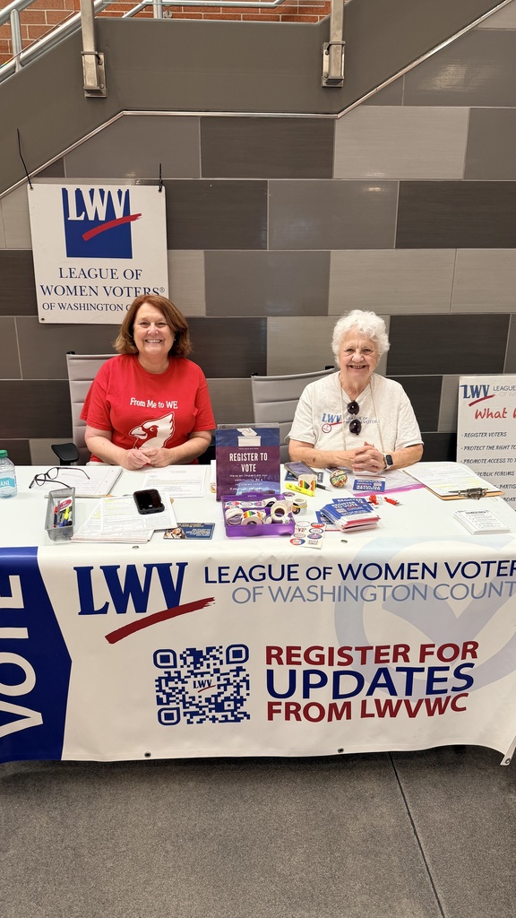 Two women at a table with a white tablecloth. One woman wears a red shirt. Behind them, a brick wall and signs read "League of Women Voters."