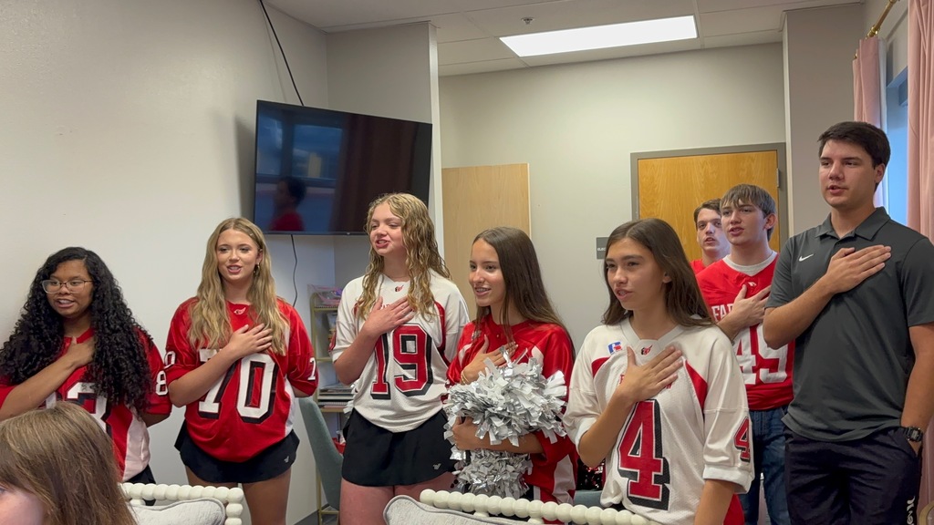 Several young athletes in red jerseys with numbers stand in a room, arms over hearts, while a coach looks on.