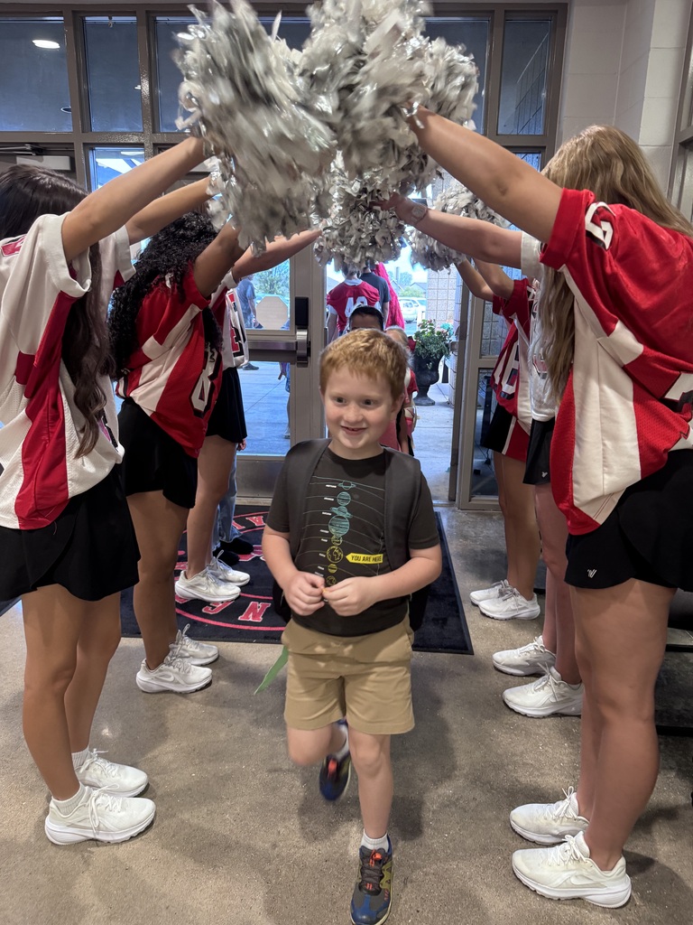 Several cheerleaders holding pom-poms over a smiling boy in a gray shirt and khaki shorts.