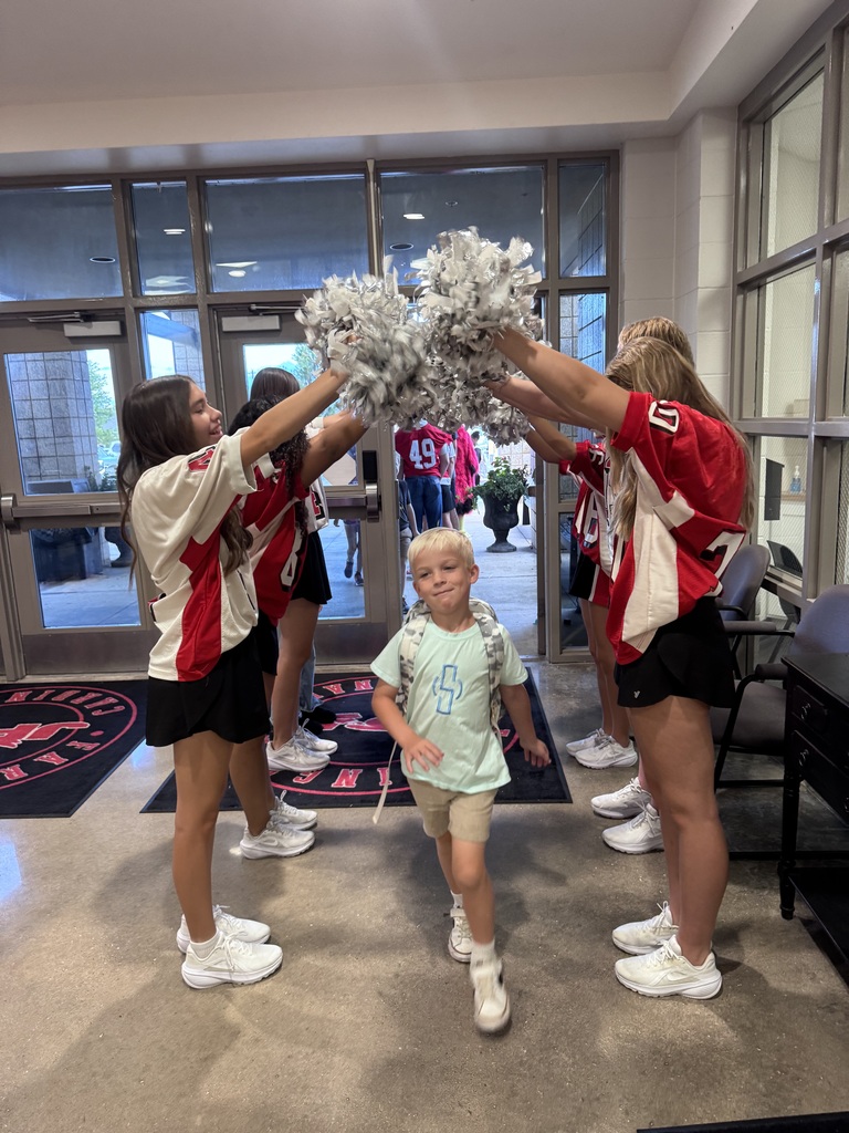Several people, including cheerleaders, stand in a room. One child walks among them. Bright sunlight filters through large glass doors.