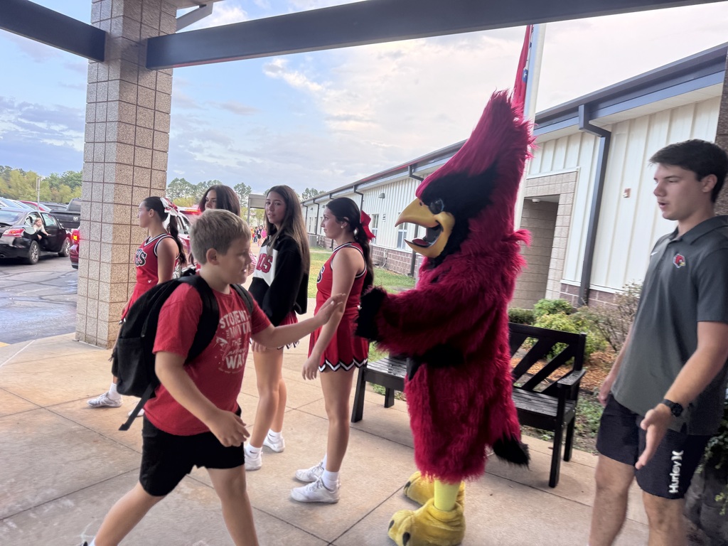 Students, one with a backpack, interact with a school mascot in front of a building.