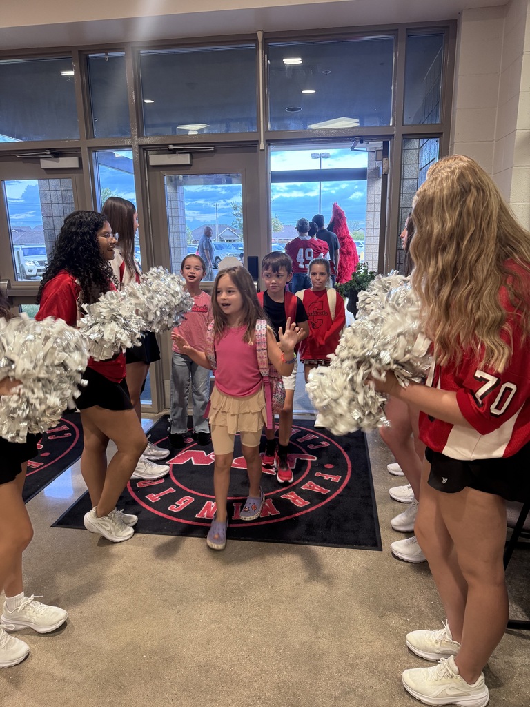 Several cheerleaders hold pom-poms in front of a glass door with a red banner. A young girl stands in the center.