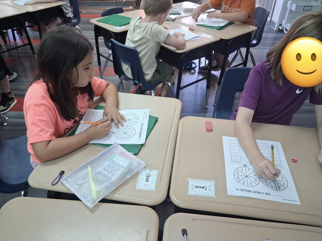 Three children, two girls and one boy, seated at desks in a classroom. Each child holds a pencil, writing on paper. A smiley face is positioned near one child.