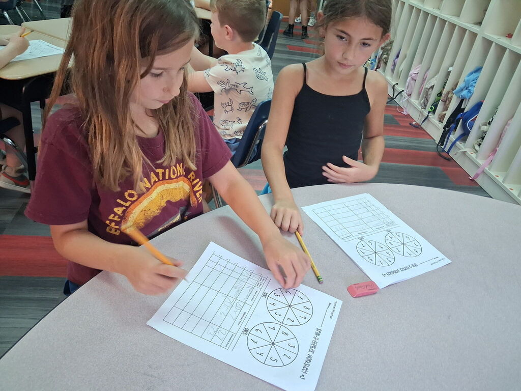 Two girls seated at a table, working on printed pages with a wheel graph and numbers.