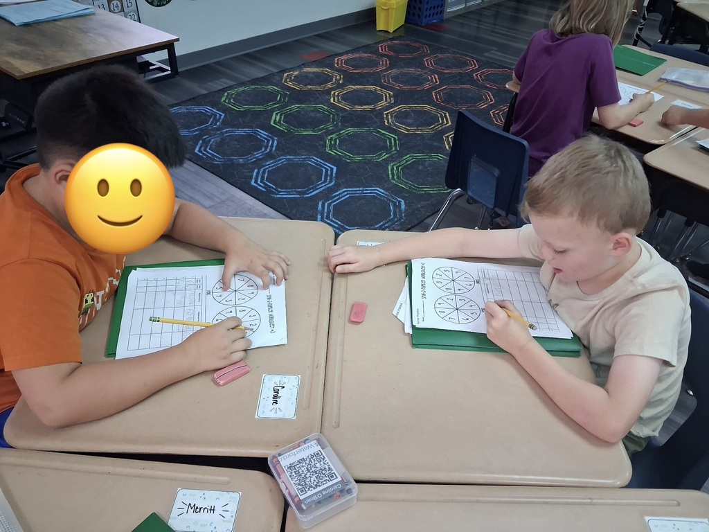 Two children working on school activities at desks. One wears an orange shirt; the other wears a white shirt.