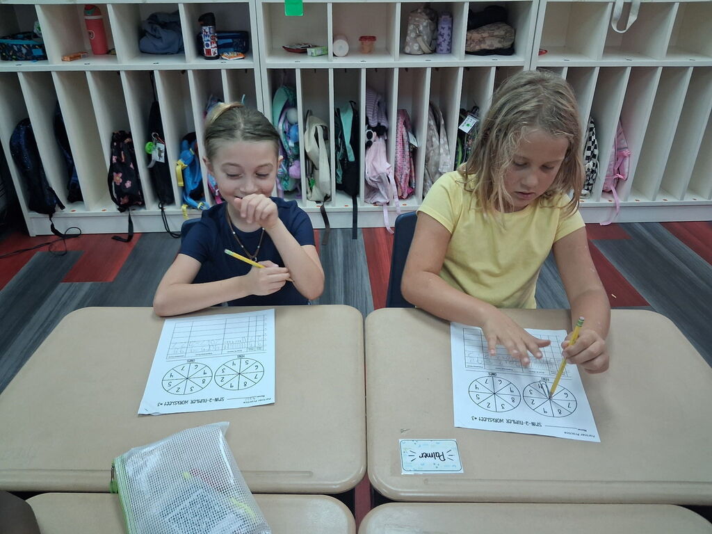 Two girls in a classroom setting, seated at desks. One uses a pencil to draw while the other watches.
