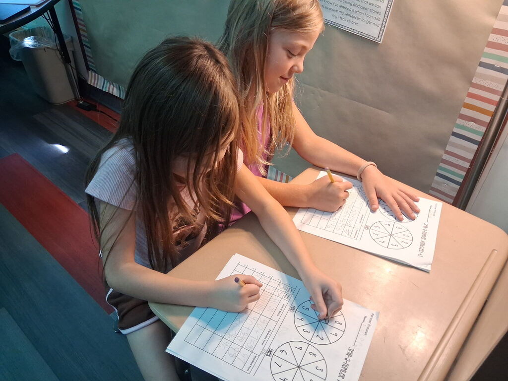 Two girls, one with long hair, the other blonde, sit at a table working on worksheets.