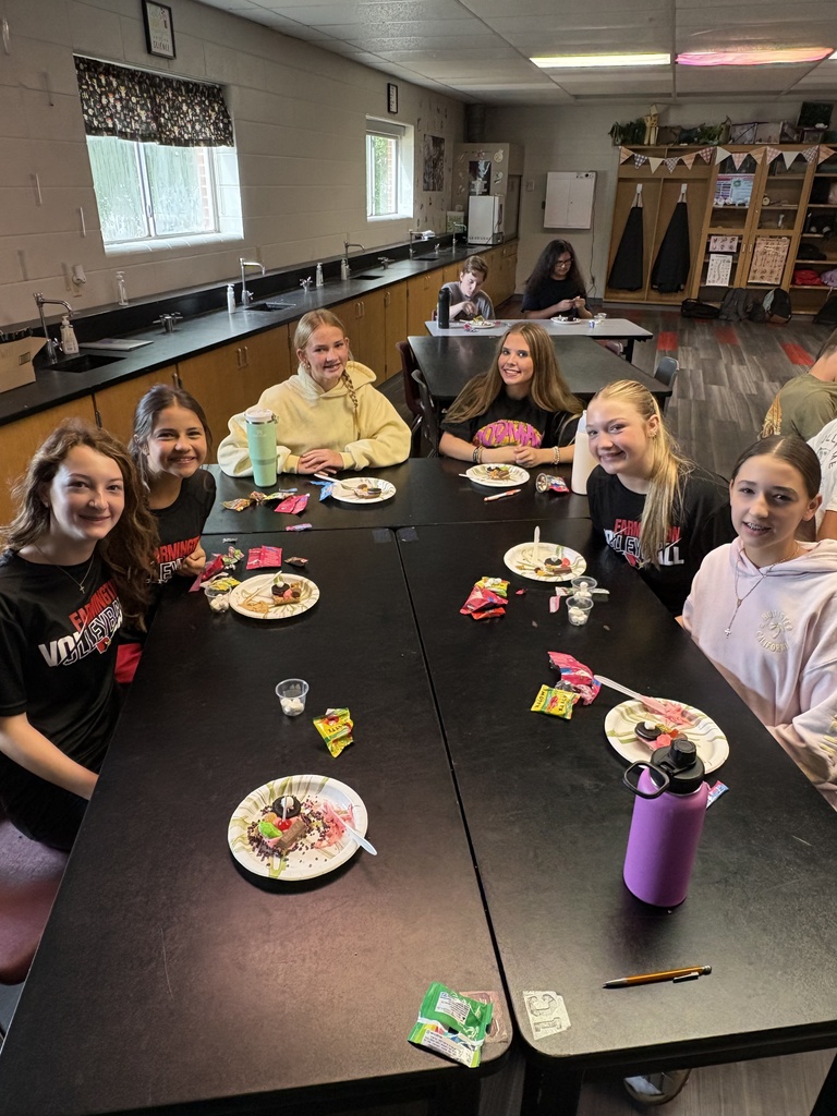 A group of students sit around tables, eating and drinking. A black table has plates of food, utensils, and a purple bottle.
