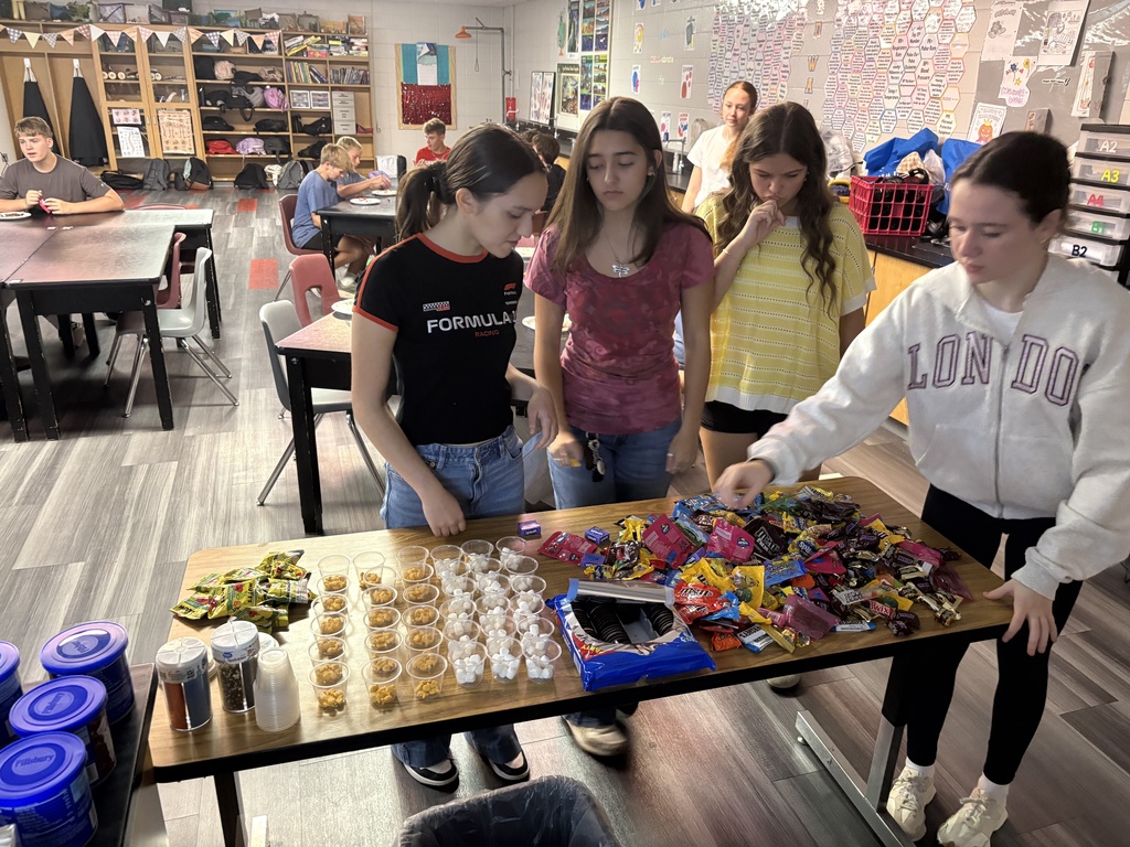Several students gather around a table filled with various candies, snacks, and other items.