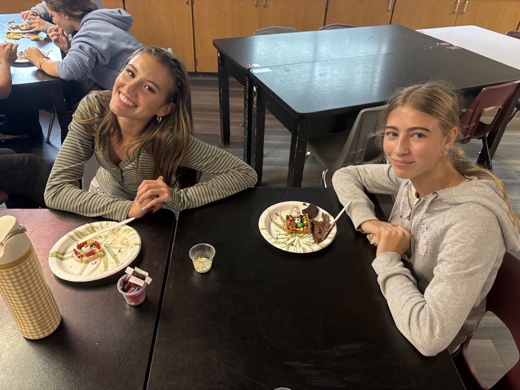 Two women sit at a table, smiling and posing. One holds a spoon, and the other holds a fork.