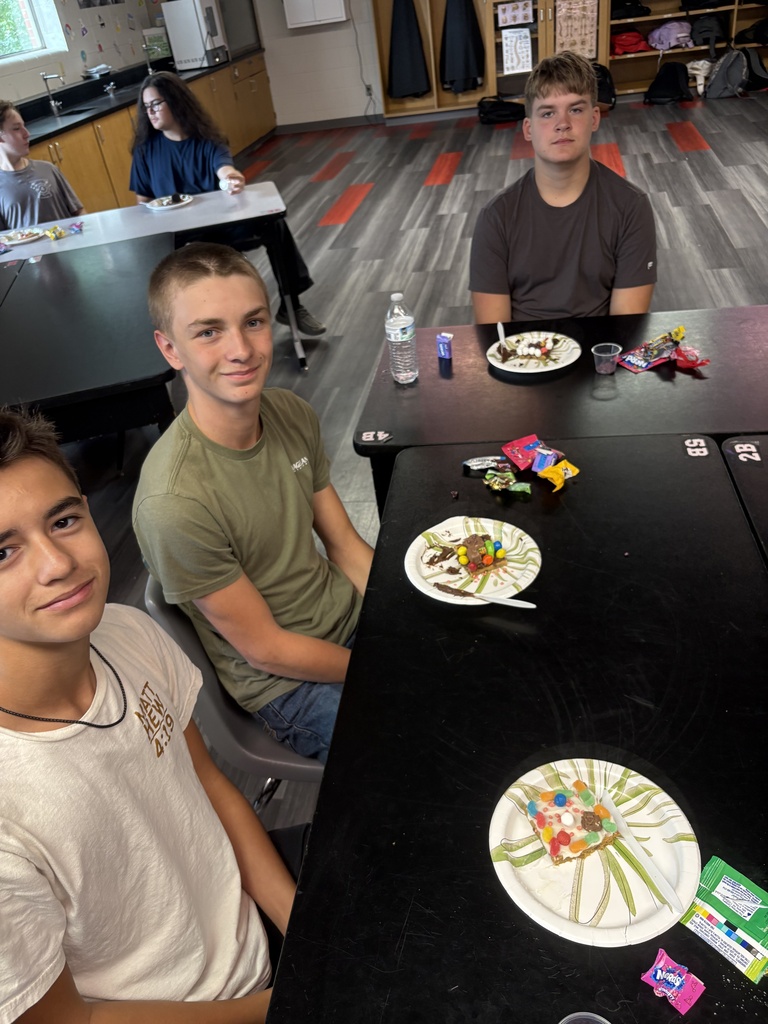 Three boys are sitting at a table, smiling. They have plates of candies on the table. Behind them, a girl is sitting at another table.