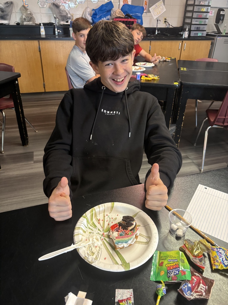 A boy with a black hoodie sits at a table, giving two thumbs up. There is a plate with a dessert in front of him.
