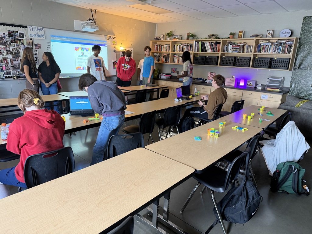 A classroom with people gathered around tables, a projector screen, and bookshelves in the background.