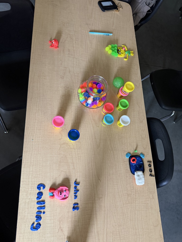 Light wood table with toys and objects. A bowl of multicolored balls is in the center. Names written on the table.