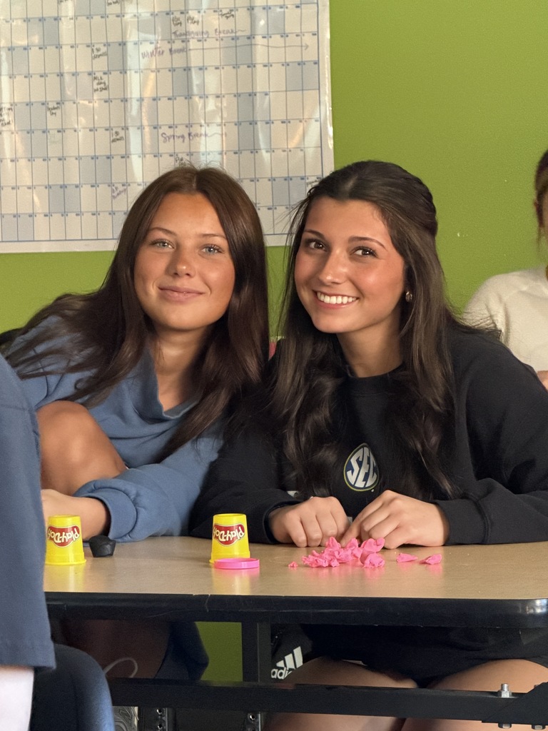 Two women sitting at a desk smile, one with a blue shirt and the other with a black shirt.