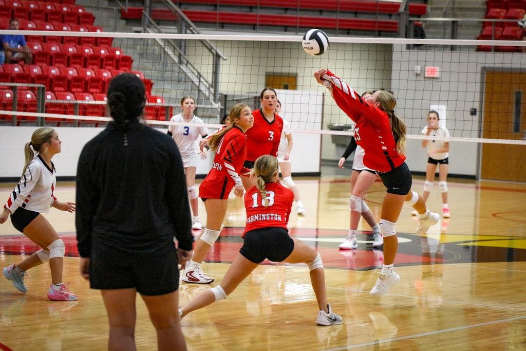 A group of female volleyball players in red and white uniforms, jumping and reaching for a ball on a court.