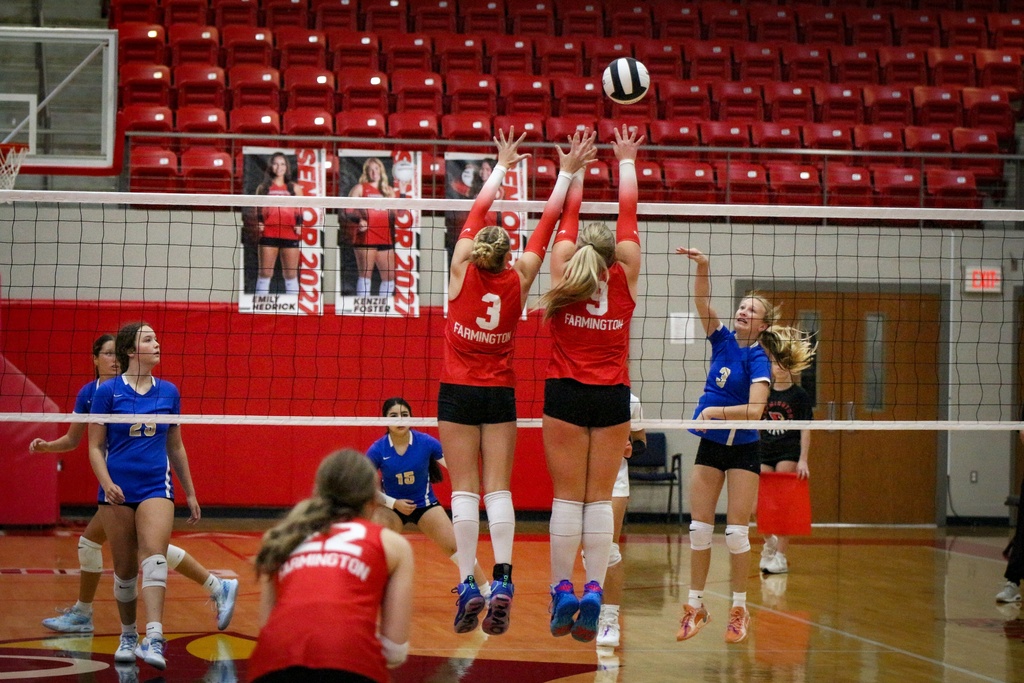 A team of female volleyball players in red and blue uniforms playing on an indoor court with a red wall.