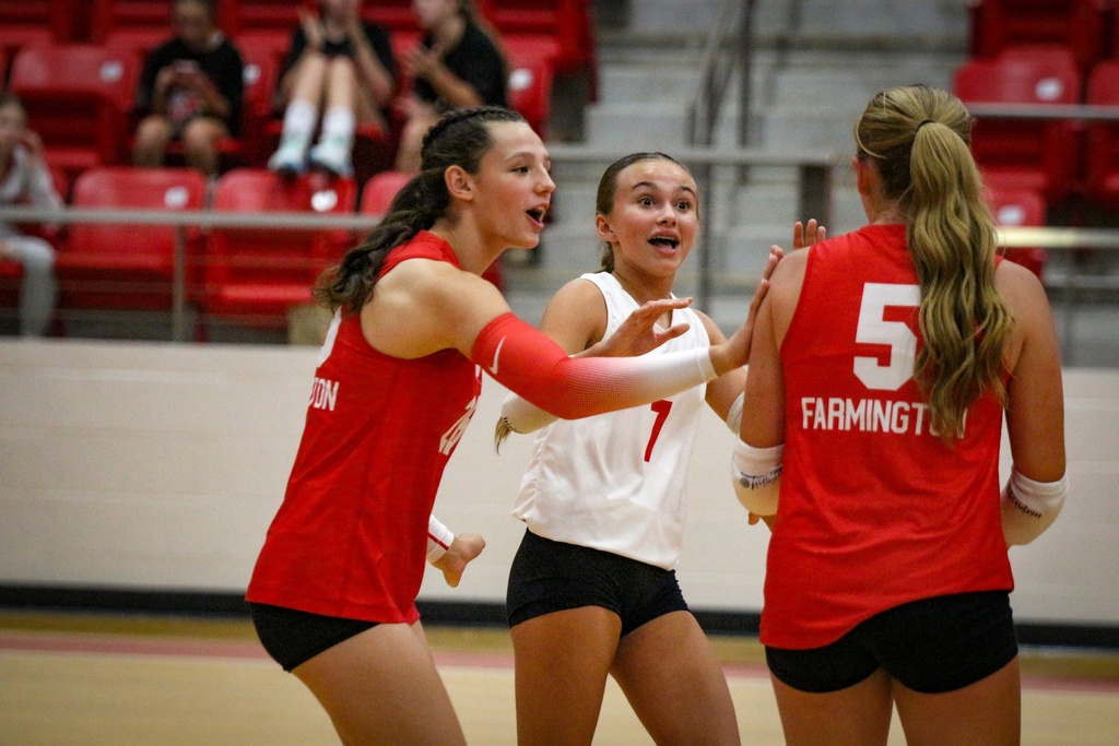 Three women celebrate on a court with a red background. Two wear red uniforms, one with number 5, and one wears a white uniform.