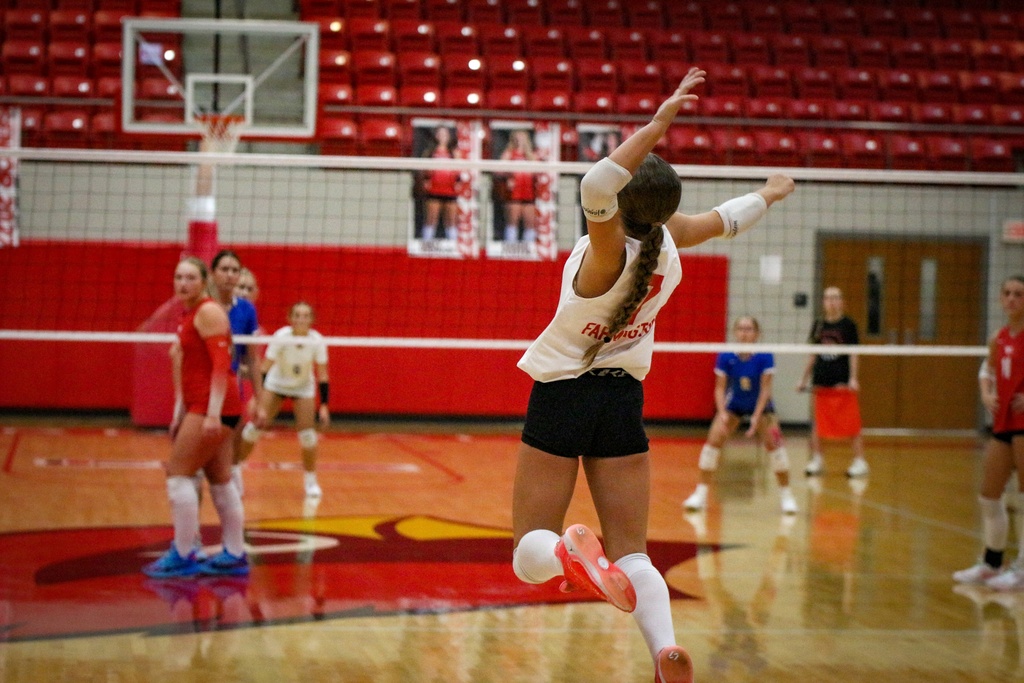 An indoor volleyball match with players in red and white uniforms. One player in white jumps to hit the ball.