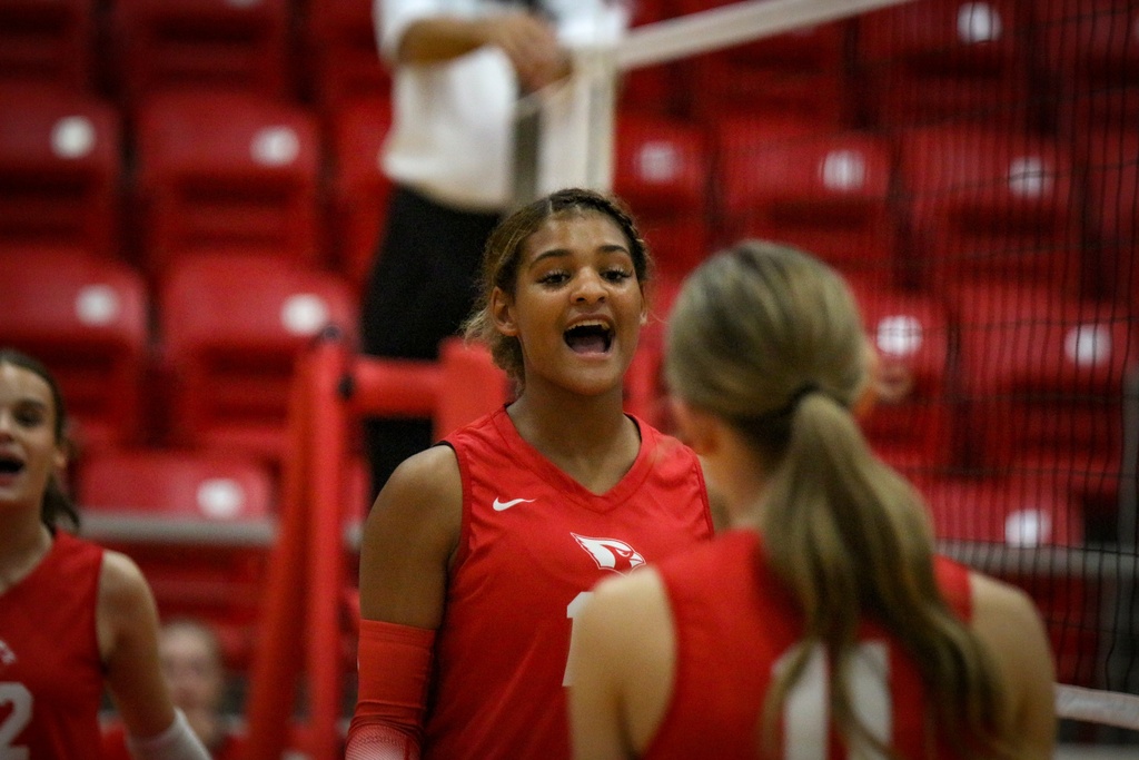A volleyball player in a red jersey stands between two teammates. All wear red uniforms. Empty red seats in the background.