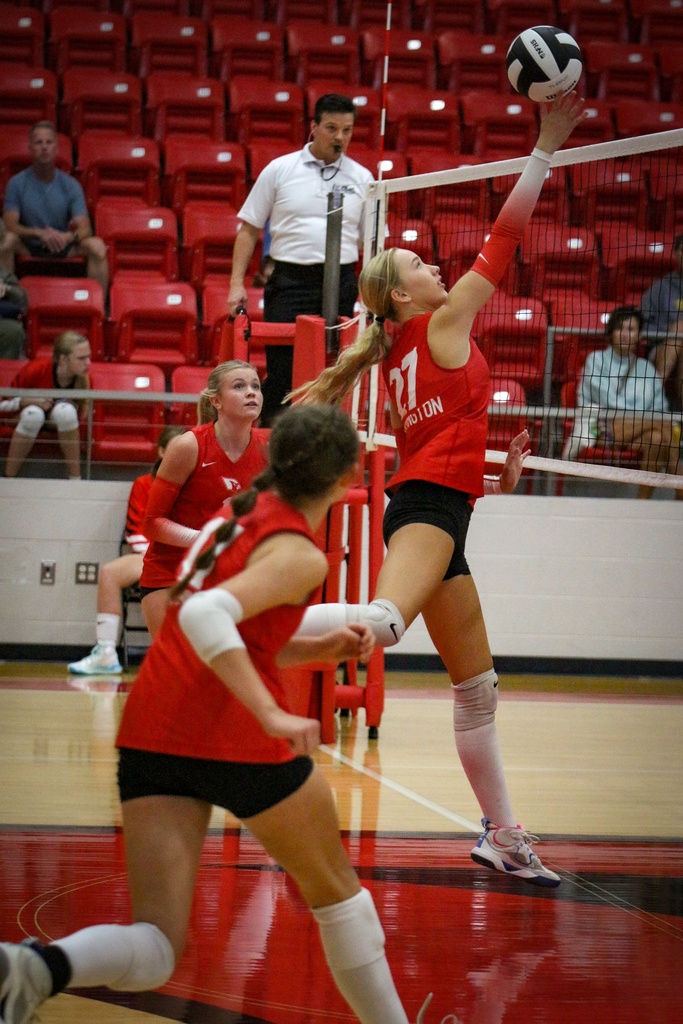 A woman in a red jersey jumps to hit a volleyball over a net. Another woman runs toward the net.