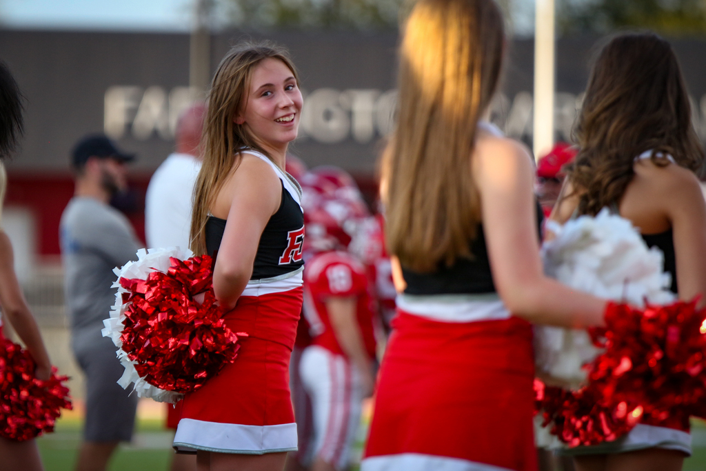 Four cheerleaders with red and white uniforms and pom-poms. A man wearing a baseball cap and a beard stands in the background.