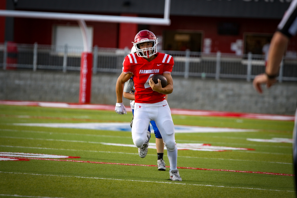 A football player in red jersey and helmet running with a ball on a field. A referee is behind.