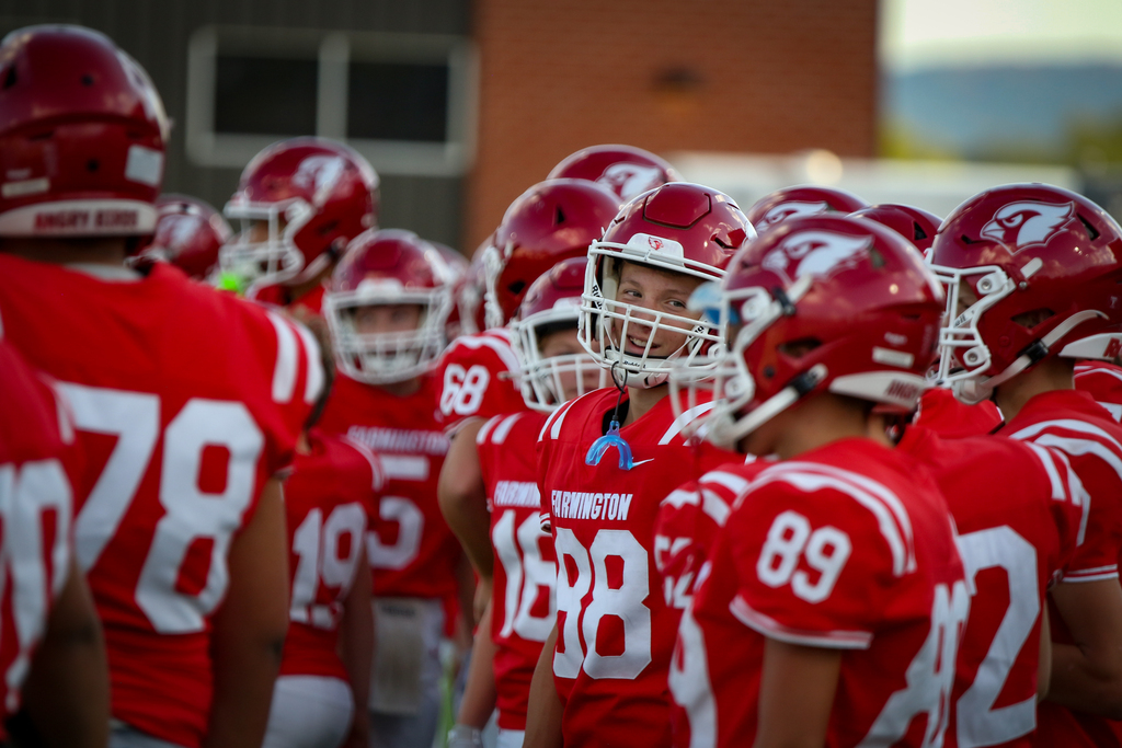 Several football players in red uniforms with helmets, numbers on back, huddle near a building.