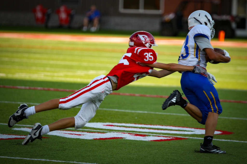 Two football players in action on a field. One in red and white, the other in blue and white.
