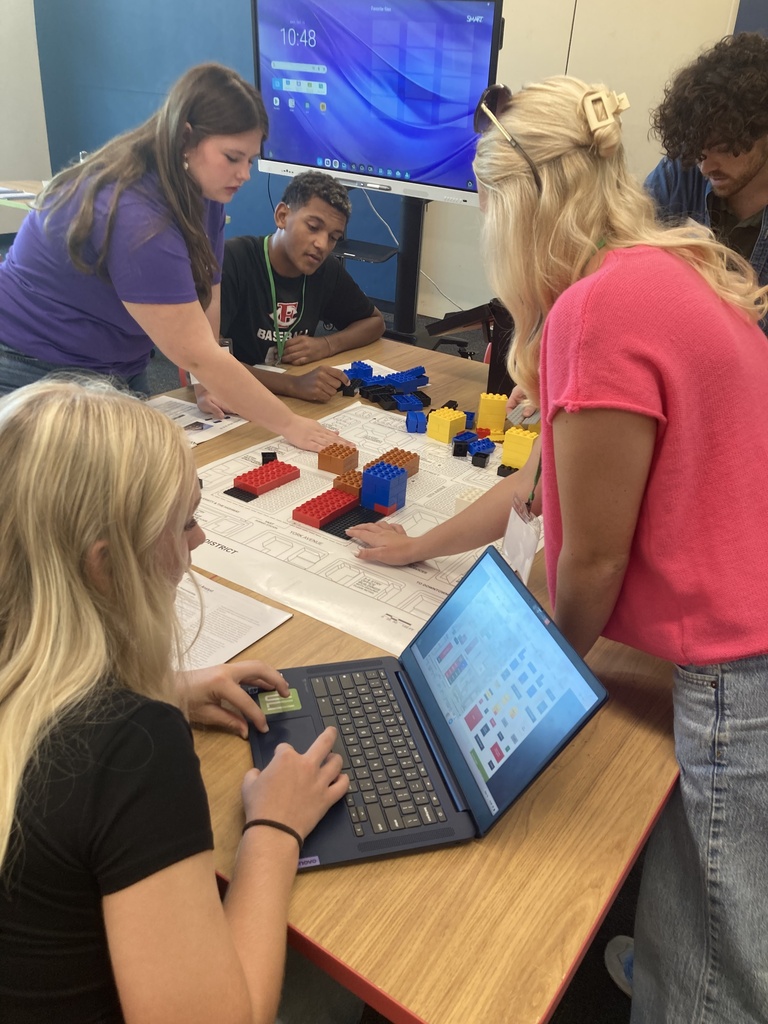 Five people, including women and men, gather around a table with a laptop and building blocks. A monitor is visible in the background.