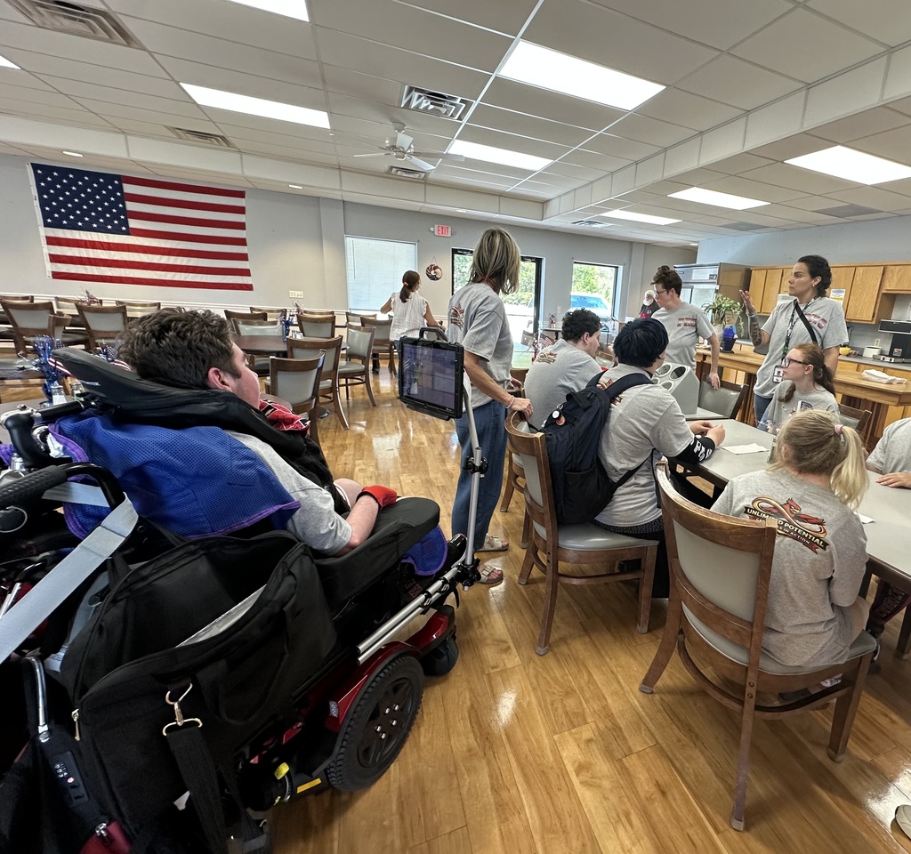 A group of people seated at tables. Some are in wheelchairs. A flag hangs from a wall.