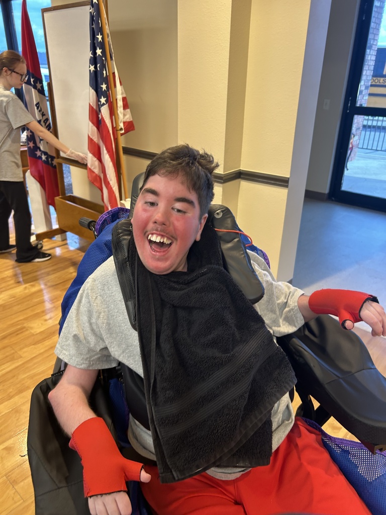 A person in a wheelchair smiles, wearing a black scarf and red arm supports. Behind them is a flag and a person standing. The room has wooden floors and a window.