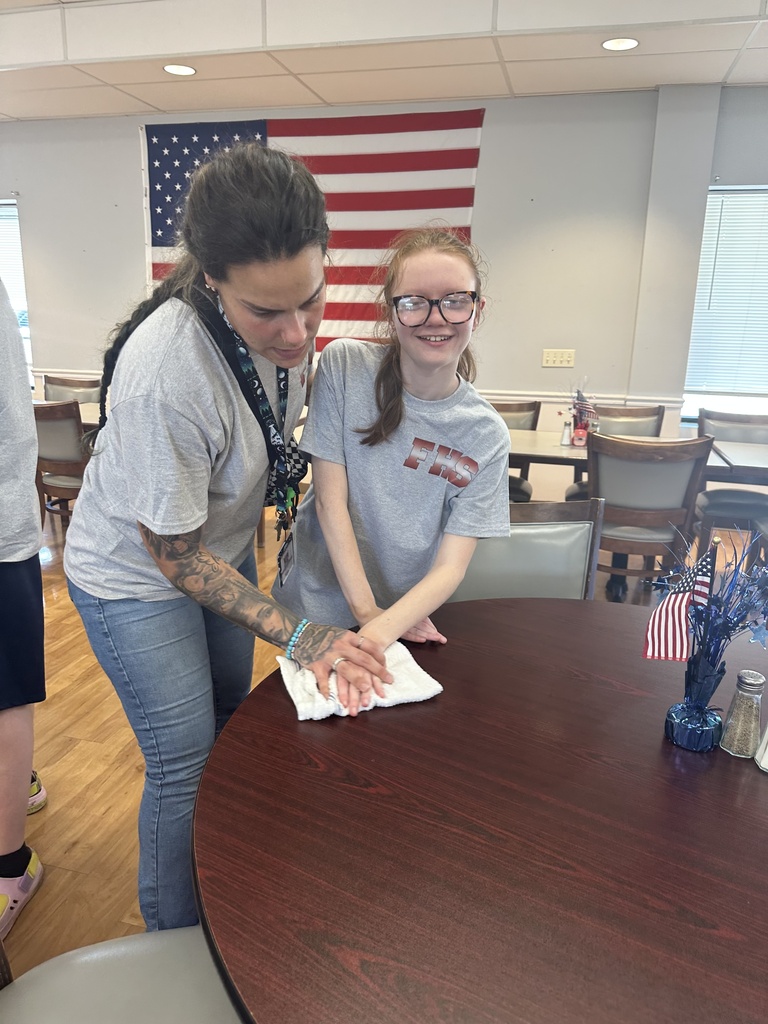 A woman and a girl are washing hands at a table with a flag on the wall.