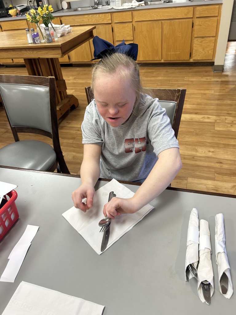 A young person with a bow in their hair folds a paper napkin. A table holds silverware.