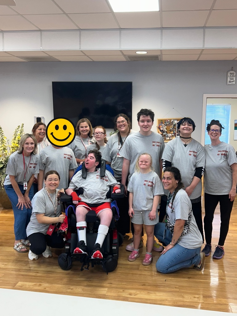 A group of people in matching gray shirts, some in wheelchairs, smiling and posing for a photo in a room.