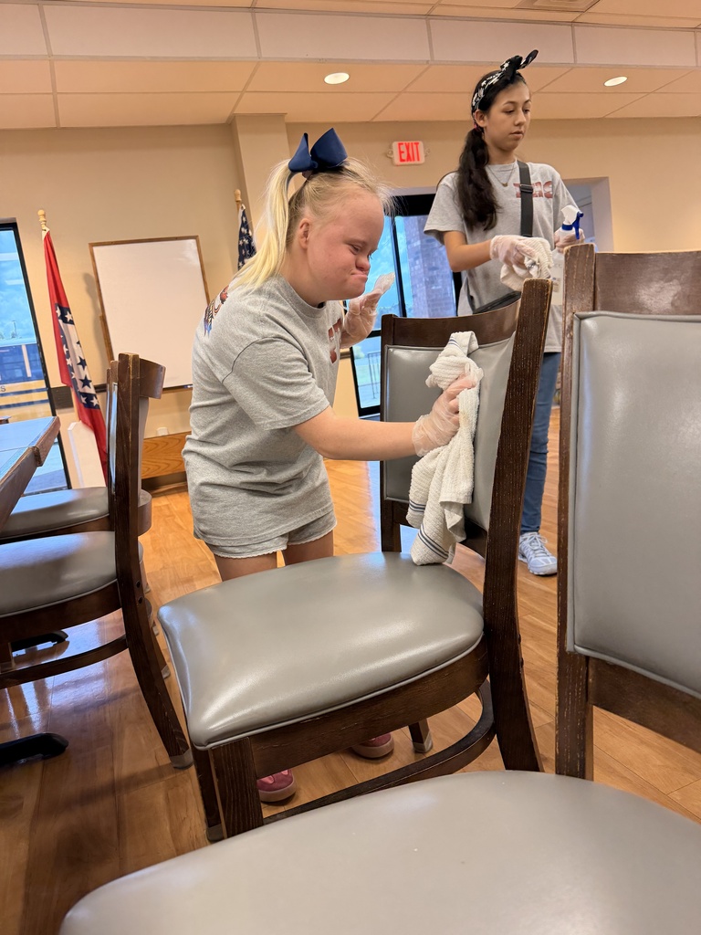 Two girls, one cleaning a chair, the other holding a cloth, in a room with wooden floors and chairs.