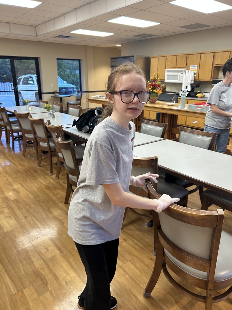 A girl in a gray shirt and glasses pushes a chair in a dining area with wooden floors.