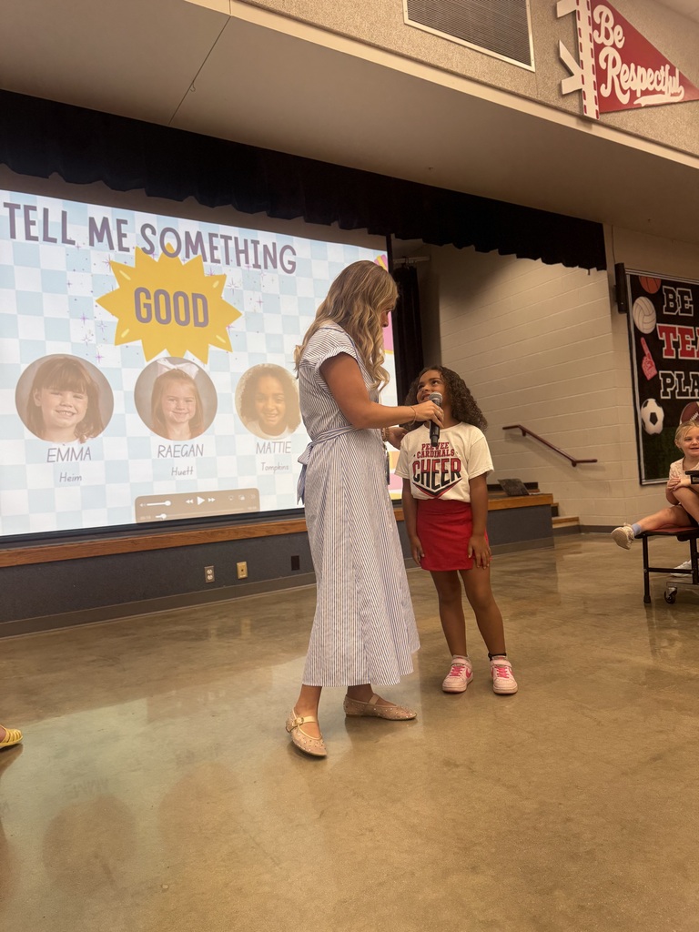 A woman holds a microphone and speaks to a young girl, both wearing summer clothing, in front of a large screen.
