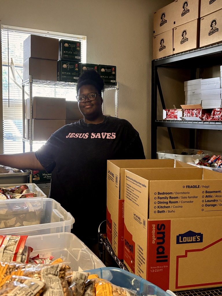 A person stands behind a table with a large box of items in a room full of boxes.