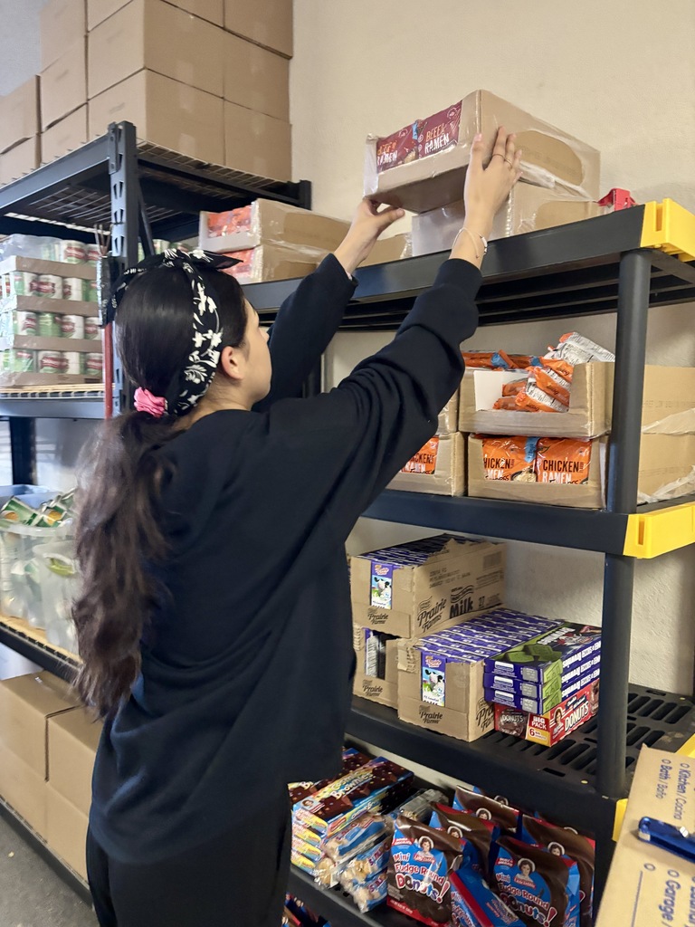 A person arranges food boxes on a shelf in a pantry. Shelves are filled with boxes and bags.